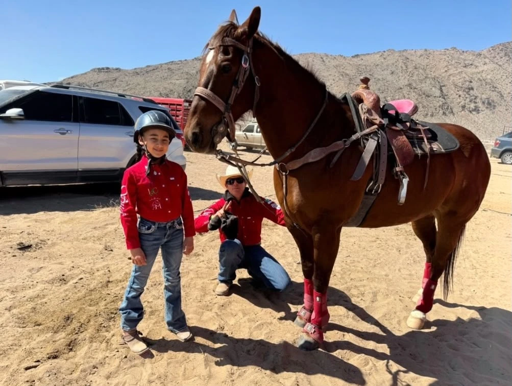una pequeña niña de 10 años esta casi lista para competir en carrera de barriles y esta junto a su caballo en el estacionamiento del ruedo junto a su padre y ambos visten una camisa roja de la marca tre