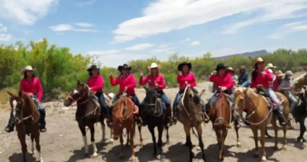 las mujeres integrantes de cabalgantes chihuahua posando para una foto montadas en sus caballos y vistiendo su blusa camisa vaquera tret color fiusha en un paisaje abierto